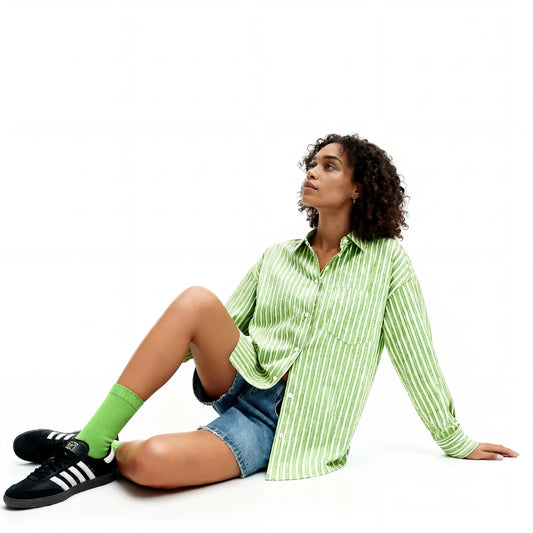 Person wearing a green striped shirt, denim shorts and green socks sitting on the floor with a white background