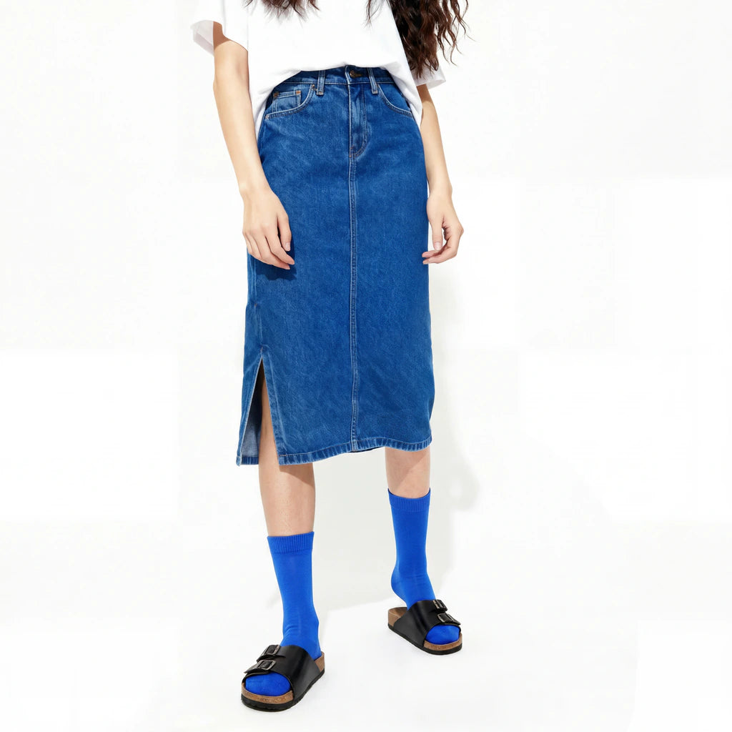 Person wearing a blue denim skirt with a white top and cobalt socks with slides on a white background