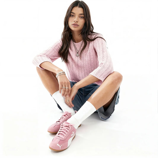 Woman wearing a pink sweater and pink sneakers with white socks sitting on a white surface.