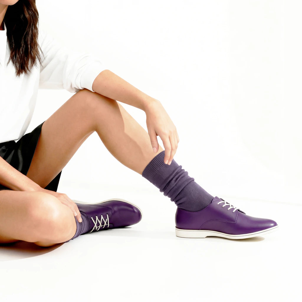 Woman sitting on the floor wearing a white sweatshirt, black shorts, and purple shoes with black socks.
