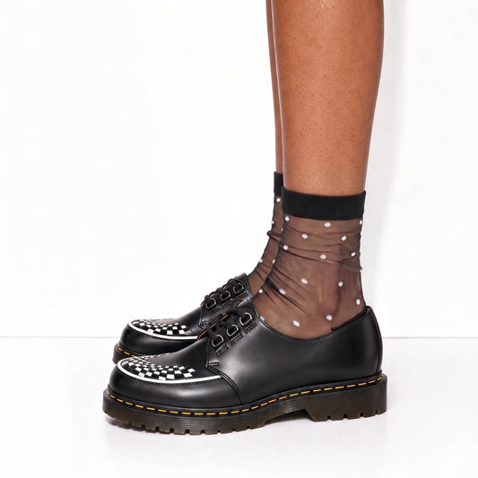 A person's lower legs wearing black leather Dr. Martens lace-up shoes with a checkerboard toe detail, paired with sheer black ankle socks featuring white polka dots, against a plain white studio background.