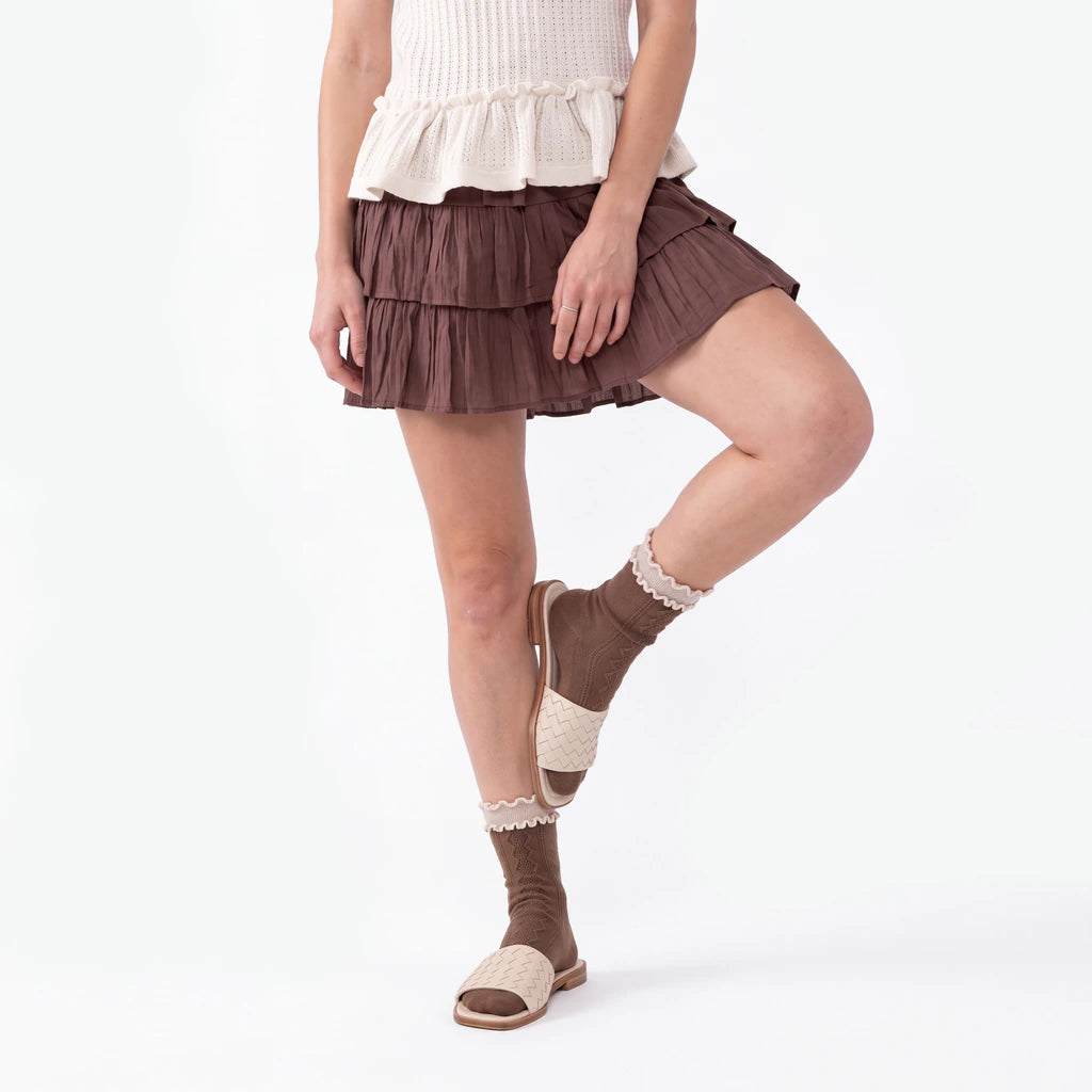 A model wearing a brown tiered ruffle mini skirt, cream knit top, brown and cream knit socks, and flat sandals, shown from the waist down against a white studio background.