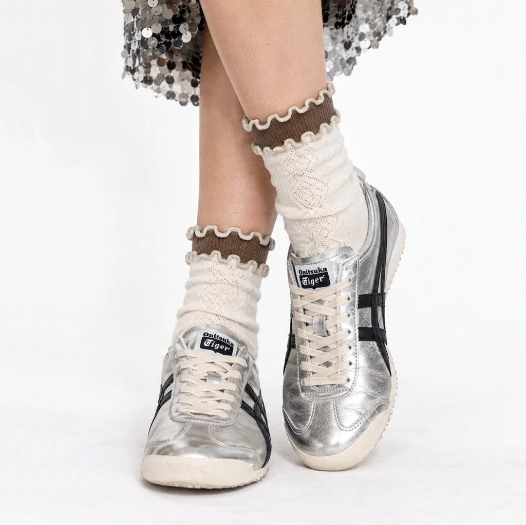 A person's legs wearing cream-colored knit socks with brown ruffled trim and silver Onitsuka Tiger sneakers, photographed against a clean white background.