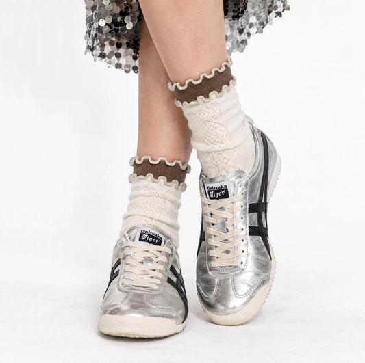 A person's legs wearing cream-colored knit socks with brown ruffled trim and silver Onitsuka Tiger sneakers, photographed against a clean white background.