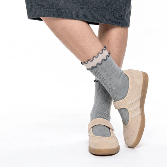 A person's legs wearing gray cable-knit socks with a ruffled trim, paired with beige Mary Jane-style shoes and a dark gray skirt, shown against a plain white background.