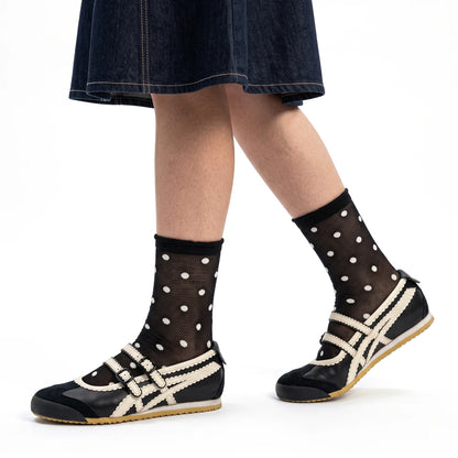 A person wearing black ankle socks with white polka dots and black slip-on shoes with cream trim, shown from the knees down against a plain white studio background.