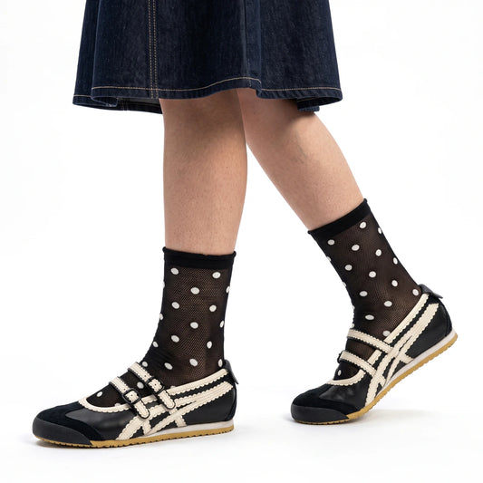 A person wearing black ankle socks with white polka dots and black slip-on shoes with cream trim, shown from the knees down against a plain white studio background.