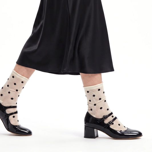 A person wearing cream mesh ankle socks with black polka dots, paired with black Mary Jane heels and a black skirt, shown against a plain white background.