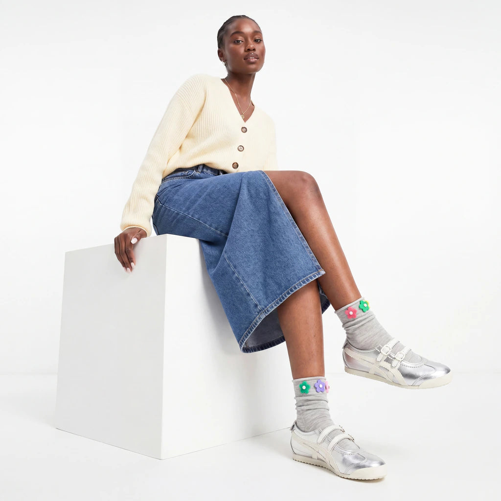 Woman sitting on a white stool wearing a cream cardigan, blue denim skirt, and colorful socks with sneakers.
