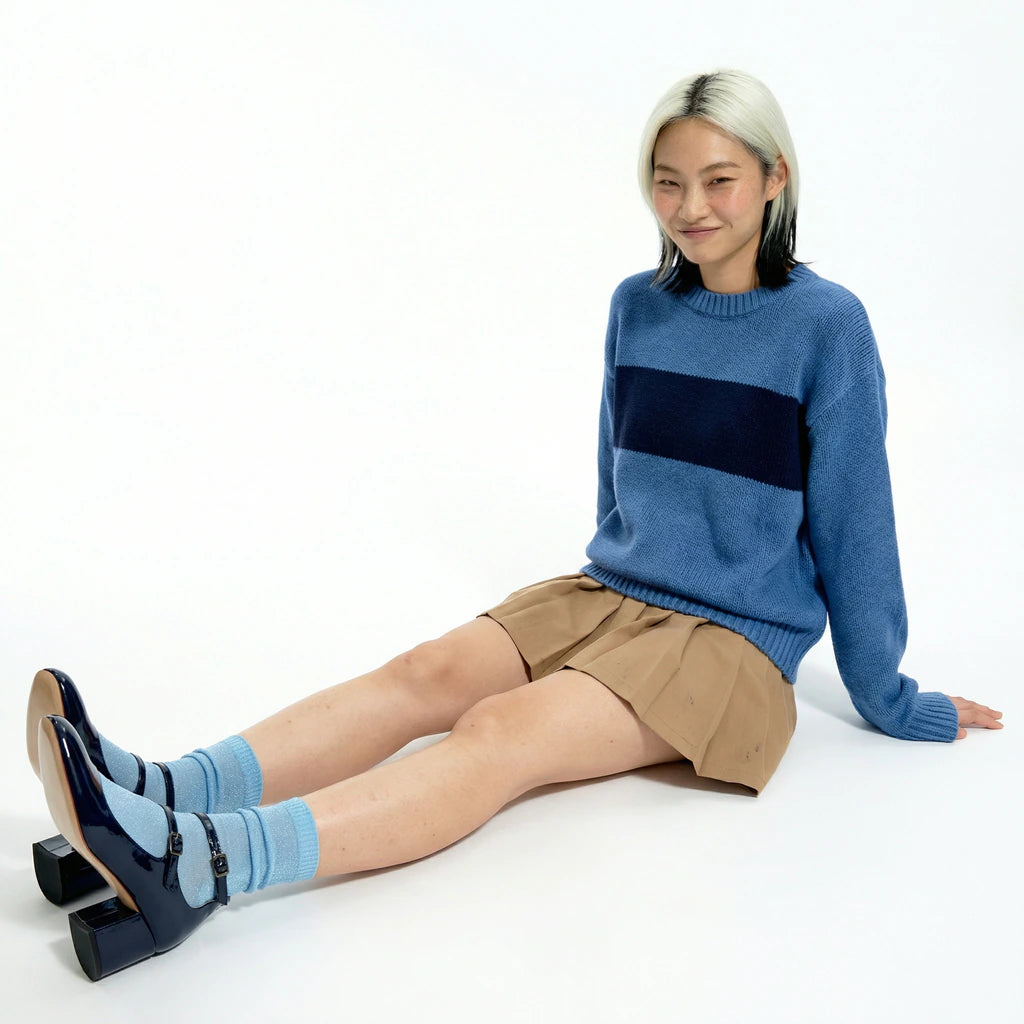 Woman wearing a blue sweater with a black stripe, beige shorts, and blue socks sitting on a white background.