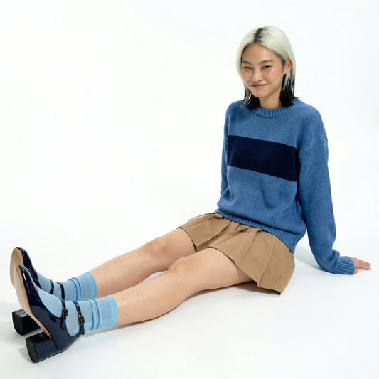 Woman wearing a blue sweater with a black stripe, beige shorts, and blue socks sitting on a white background.