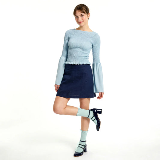A woman modeling a light blue long-sleeve textured knit top with flared bell sleeves, paired with a dark navy mini skirt, light blue socks, and dark navy Mary Jane heels, against a solid white background.