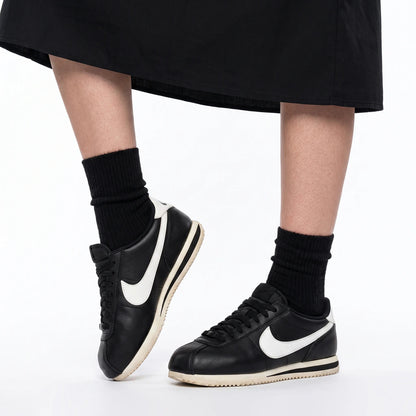 A person wearing black Nike Cortez sneakers with white Swooshes, paired with black crew socks and a black skirt, shown from the knees down against a white studio background.