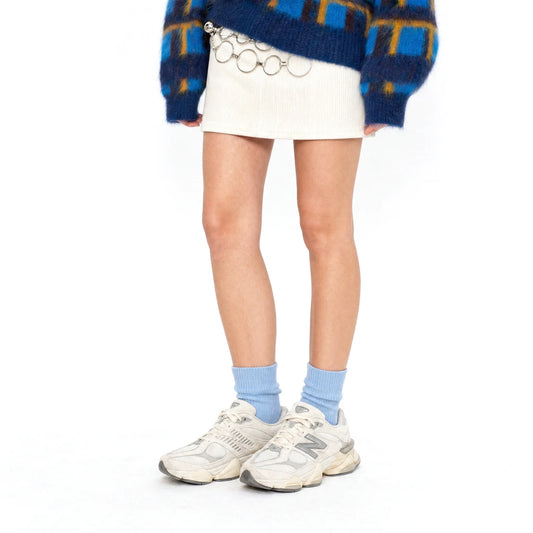 A person from the waist down wearing a cream mini skirt with a silver chain detail, a blue and yellow patterned knit sweater, light blue socks, and New Balance sneakers on a white background.