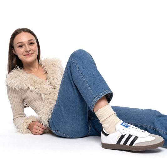 A woman reclining on a white studio floor wearing a cream ribbed sweater with a fluffy collar, blue jeans, cream socks, and white Adidas Samba sneakers with black stripes.