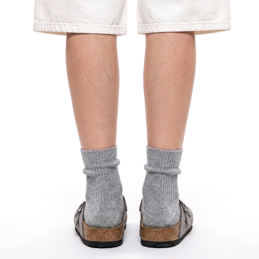 Rear view of a person wearing light gray ribbed crew socks and brown leather slip-on clog-style shoes with cork midsoles, against a plain white background.