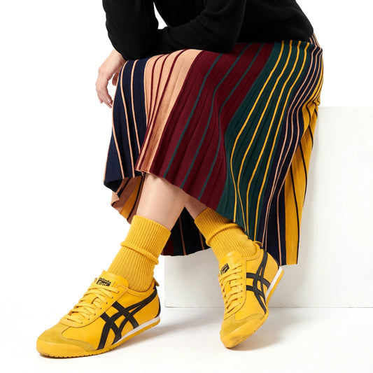 A person seated on a white stool wearing a pleated striped midi skirt in red, navy, green, yellow, and beige tones, paired with bright yellow socks and yellow Onitsuka Tiger sneakers against a white studio background.