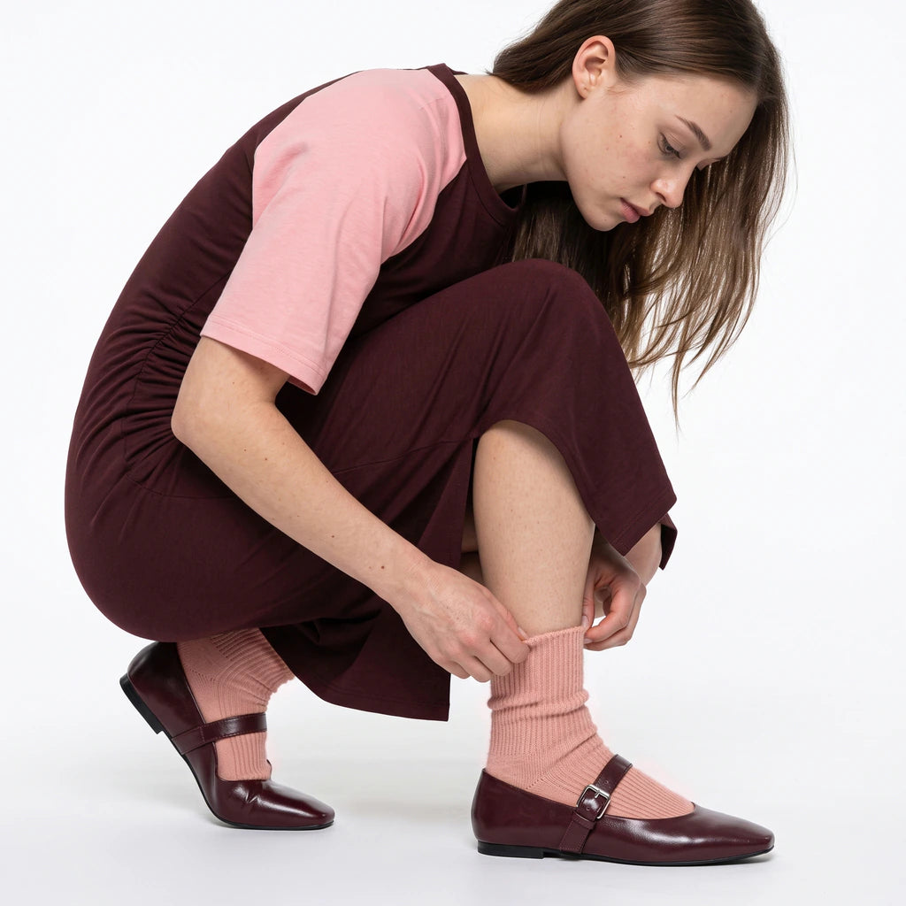A woman crouches on a white studio background, adjusting a ribbed light pink sock while wearing a dark burgundy dress with a light pink short-sleeve top, matching burgundy flats, and the same pink socks.