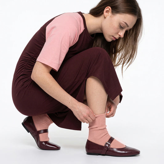 A woman crouches on a white studio background, adjusting a ribbed light pink sock while wearing a dark burgundy dress with a light pink short-sleeve top, matching burgundy flats, and the same pink socks.