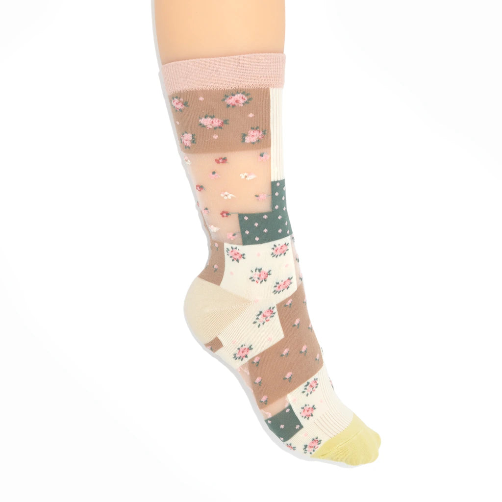 A single knee-high patchwork sock displayed on a leg against a plain white background. The sock features a patchwork design with floral prints in pink and green on beige and cream panels, a solid pink cuff, and a pale yellow toe.