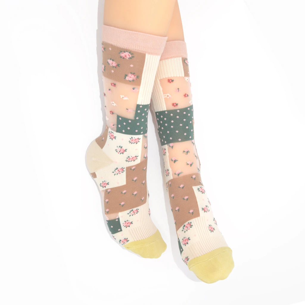 A pair of knee-high socks with a patchwork design featuring floral prints, polka dots, and solid color blocks in beige, pink, green, and cream, worn on a model's legs against a white background.