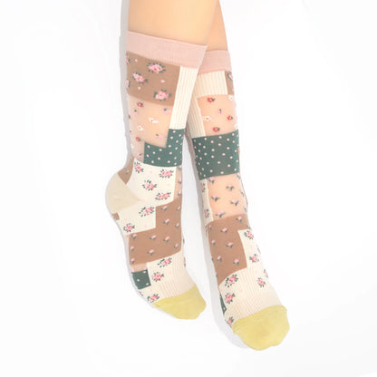 A pair of knee-high socks with a patchwork design featuring floral prints, polka dots, and solid color blocks in beige, pink, green, and cream, worn on a model's legs against a white background.