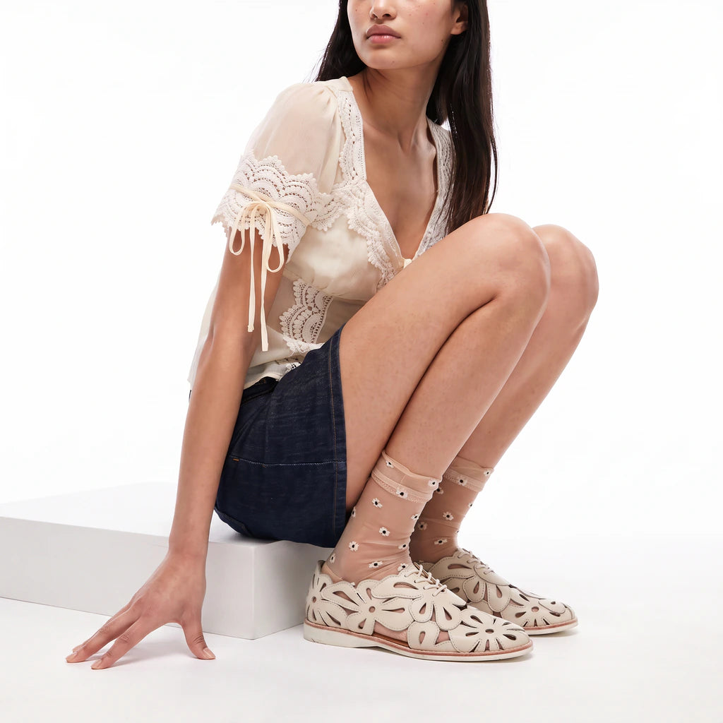 A model wearing a cream lace-trim blouse, dark denim shorts, and beige cutout lace-up flat sandals with sheer socks, seated on a white block against a white studio background.