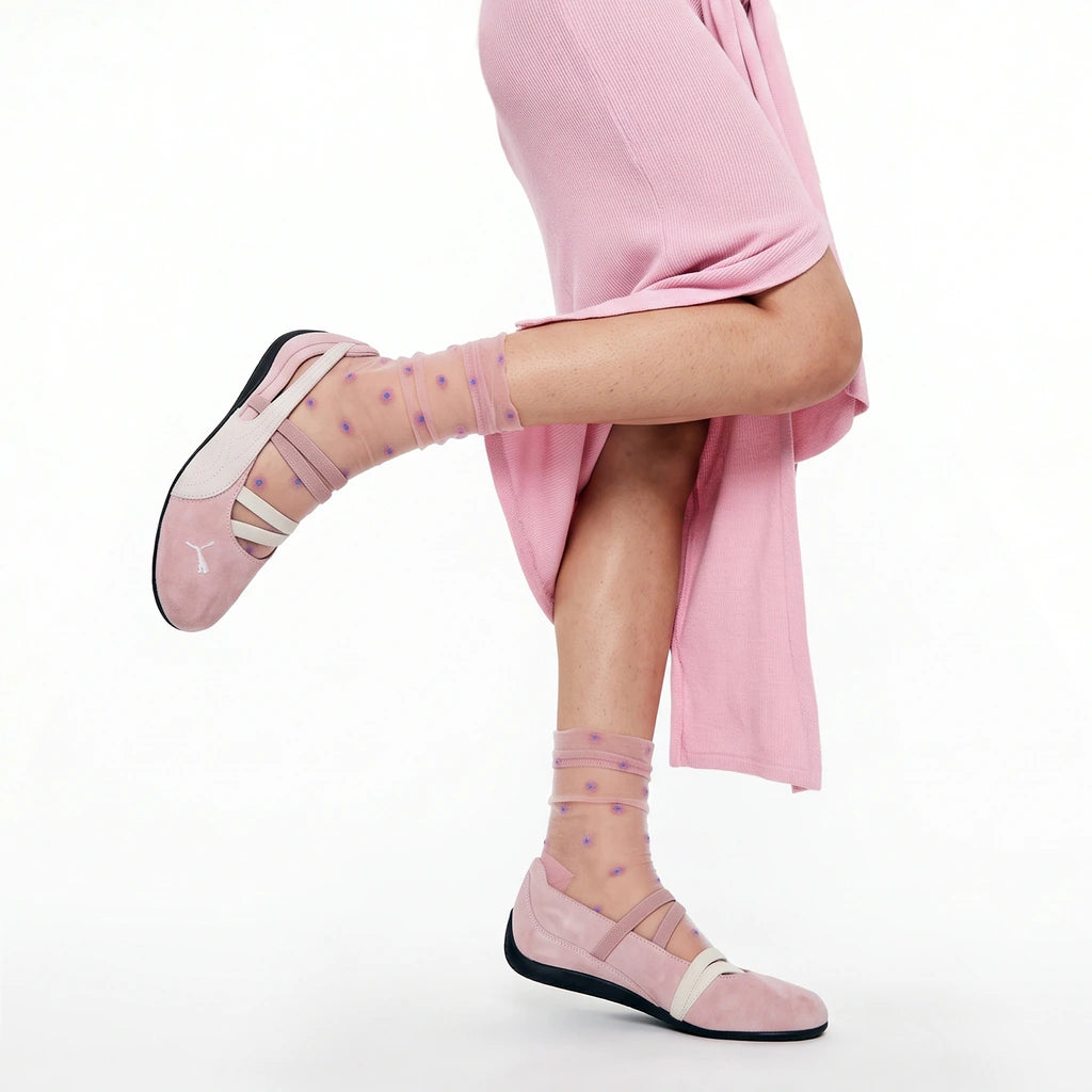 A person wearing pink ballet flats with sheer pink socks dotted with small purple stars, posed against a clean white studio background.