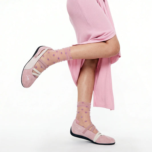 A person wearing pink ballet flats with sheer pink socks dotted with small purple stars, posed against a clean white studio background.