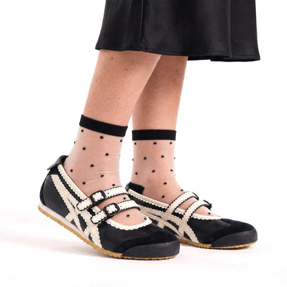 Model wearing sheer black polka dot quarter crew socks with a black ribbed cuff, styled with black and cream Mary Jane sneakers and a black satin midi skirt.