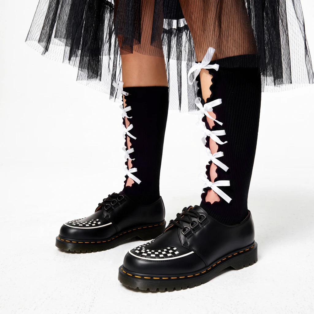 Black knee-high ribbed socks with scalloped front cut-outs and white ribbon bows, styled with black checkerboard platform shoes and a layered black tulle skirt.