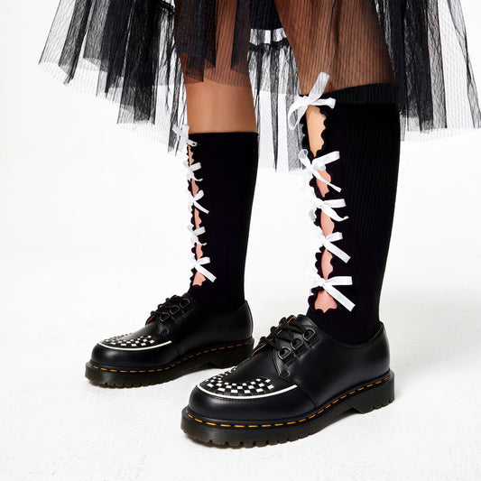 Black knee-high ribbed socks with scalloped front cut-outs and white ribbon bows, styled with black checkerboard platform shoes and a layered black tulle skirt.