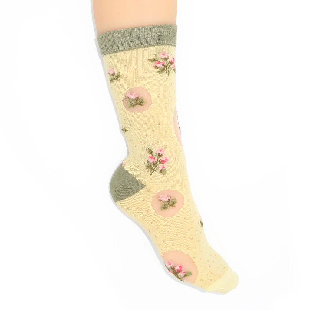 A single ankle sock displayed on a foot against a plain white background. The sock has a pale yellow base with tiny pink polka dots, pink circular motifs containing small flowers, and solid olive green ribbed cuffs, heels, and toes.