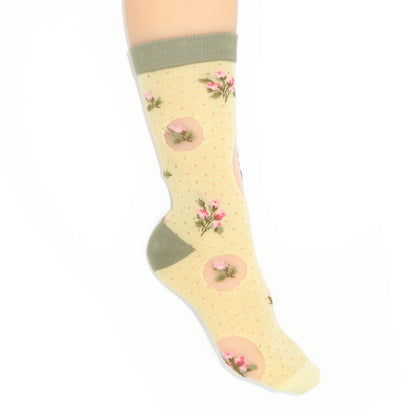 A single ankle sock displayed on a foot against a plain white background. The sock has a pale yellow base with tiny pink polka dots, pink circular motifs containing small flowers, and solid olive green ribbed cuffs, heels, and toes.