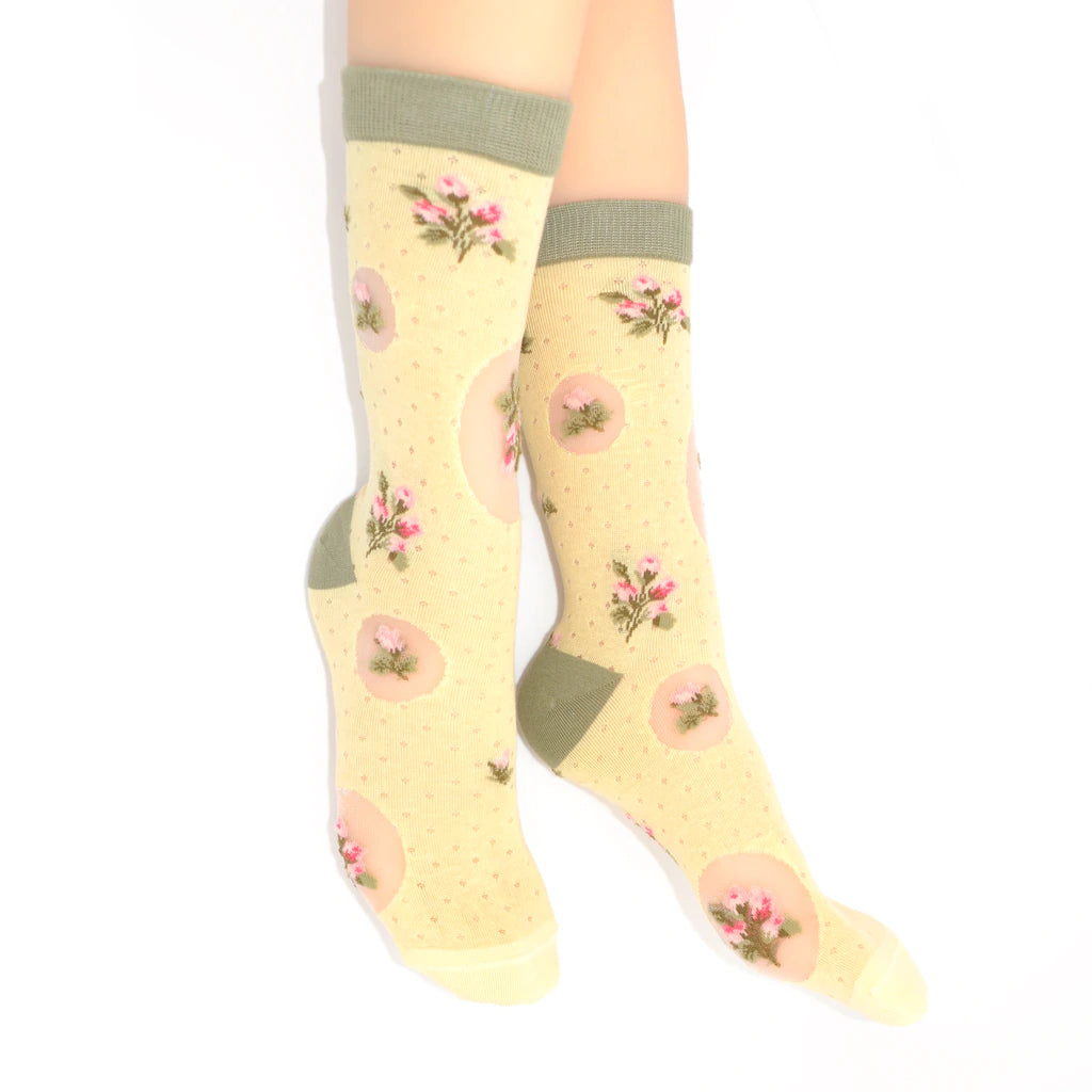 A pair of knee-high socks worn on a model's legs, featuring a pale yellow base with pink floral motifs, pink oval patches, and sage green cuffs and heels.