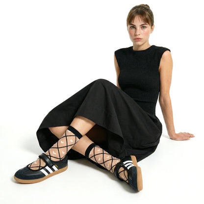 model sitting cross legged wearing a black shirred bodice dress and sheer heart criss cross socks in black contrast and black adidas samba janes