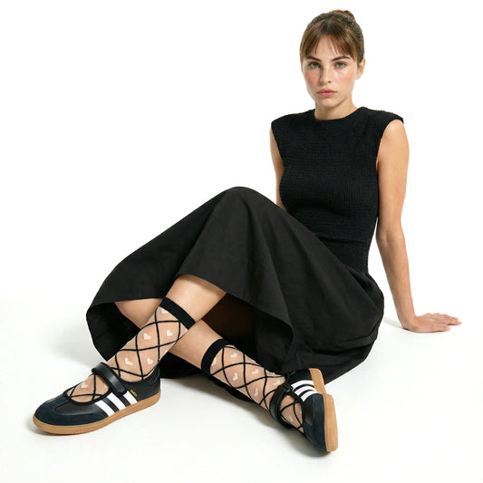 model sitting cross legged wearing a black shirred bodice dress and sheer heart criss cross socks in black contrast and black adidas samba janes