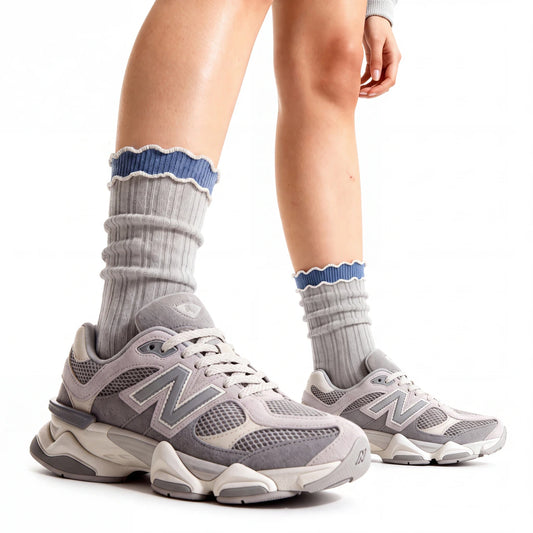 A person wearing gray New Balance sneakers with gray ribbed socks featuring a blue scalloped trim, shown against a clean white background.