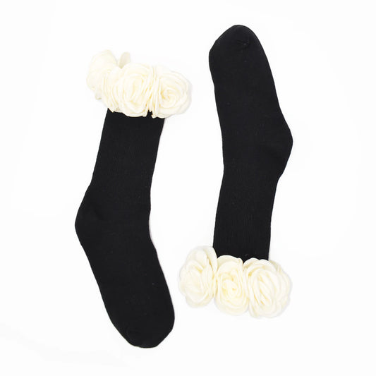 A pair of black crew socks with large white fabric flower embellishments at the top and bottom cuffs, displayed on a plain white background.