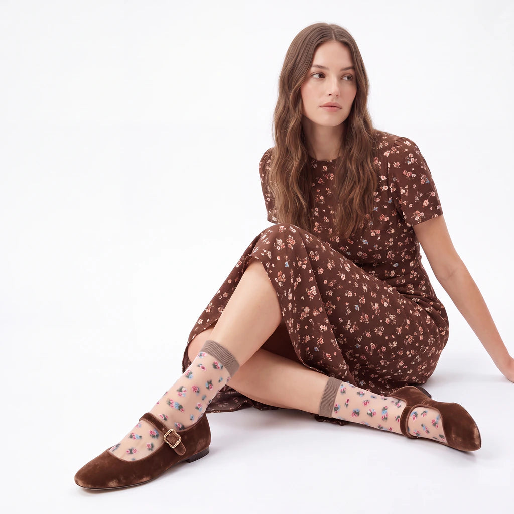 A woman sits on a white studio floor wearing a brown short-sleeve dress with a small floral print, sheer patterned socks, and brown Mary Jane flats.